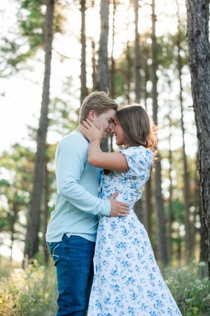 Romantic couple embraces in a golden sunlit forest, with woman wearing blue floral dress and man in light blue shirt.