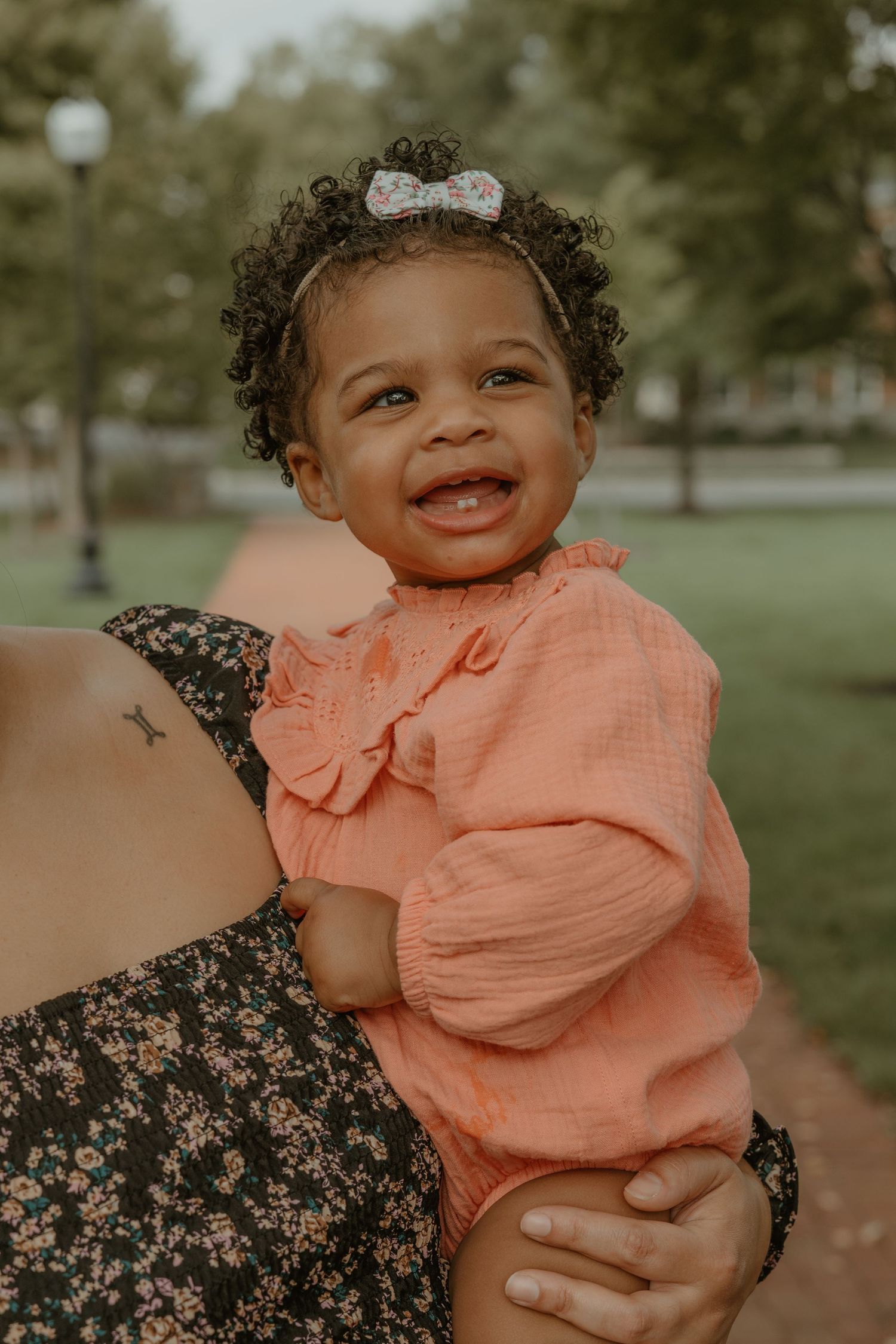 Toddler wearing coral colored outfit smiling brightly outdoors in park setting.