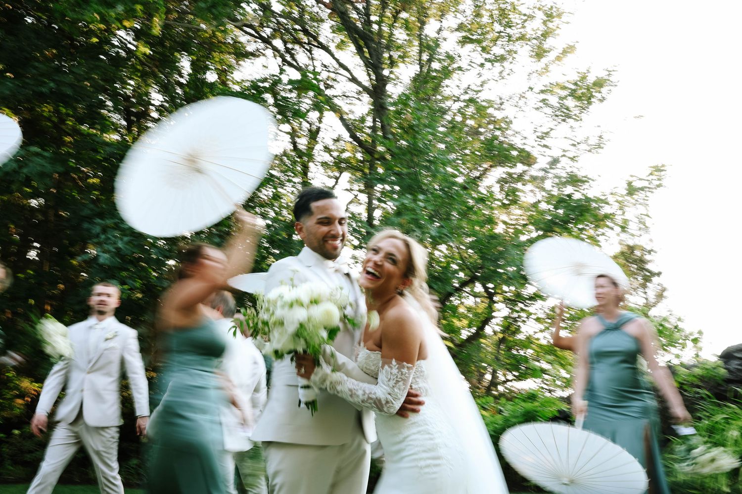 Wedding party celebrates outdoors under green trees on a sunny day.