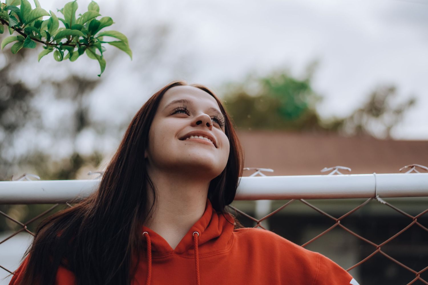 Woman in orange hoodie smiling against a chain link fence.