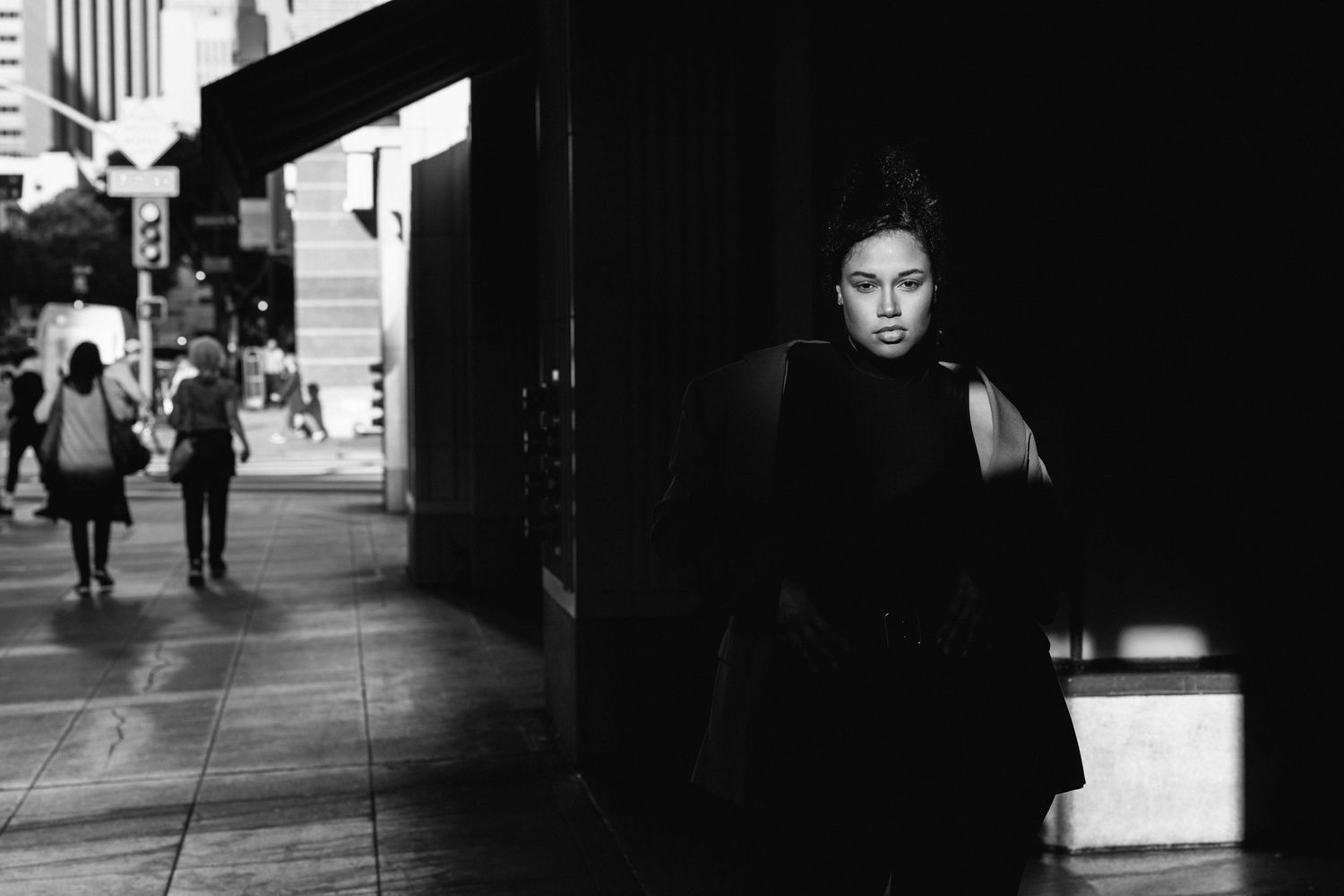 A dramatic black and white street photograph with strong shadows and contrast showing someone in dark clothing on a city sidewalk.