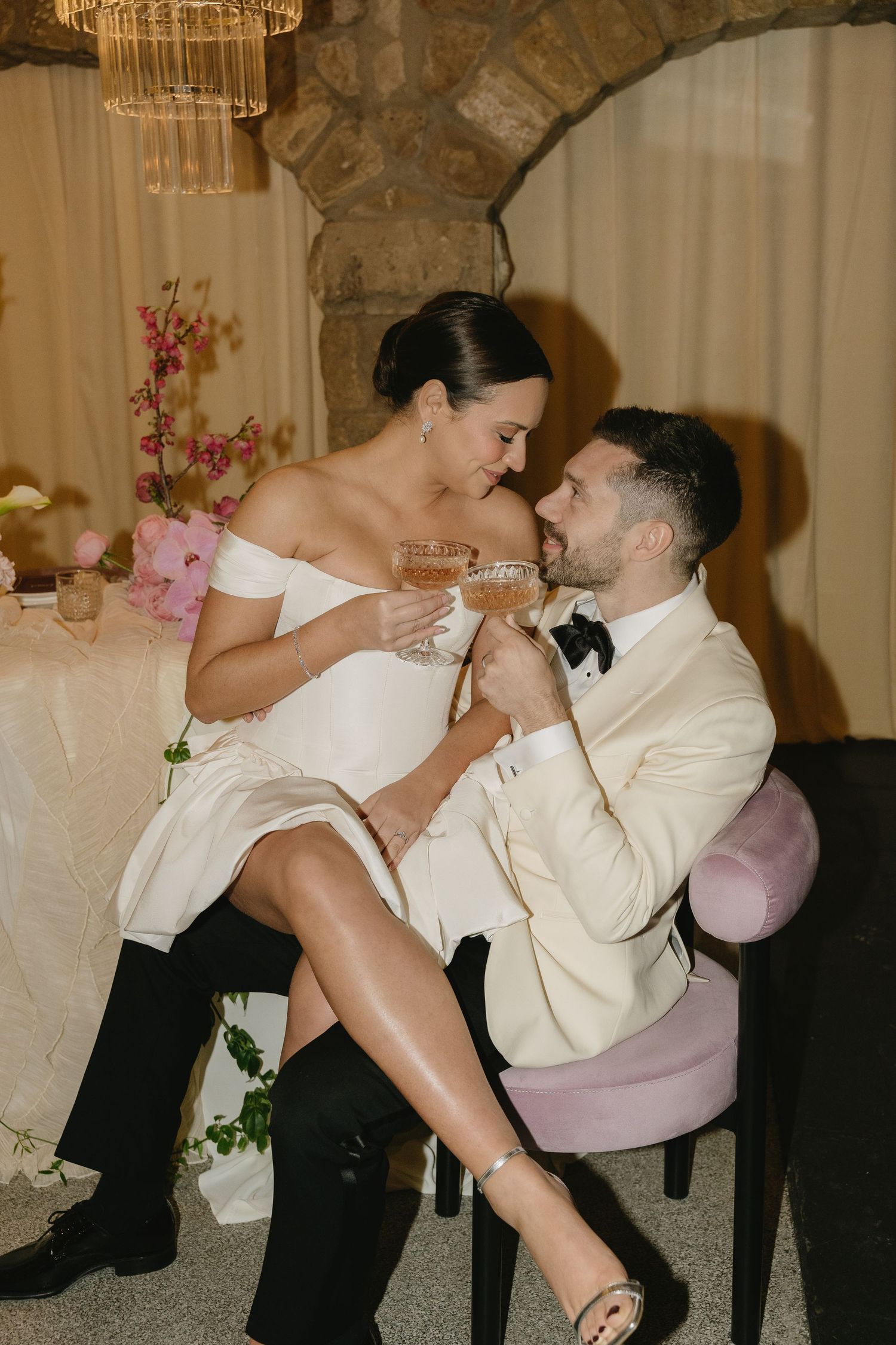 Playful couple in formal attire sharing intimate moment under crystal chandelier.