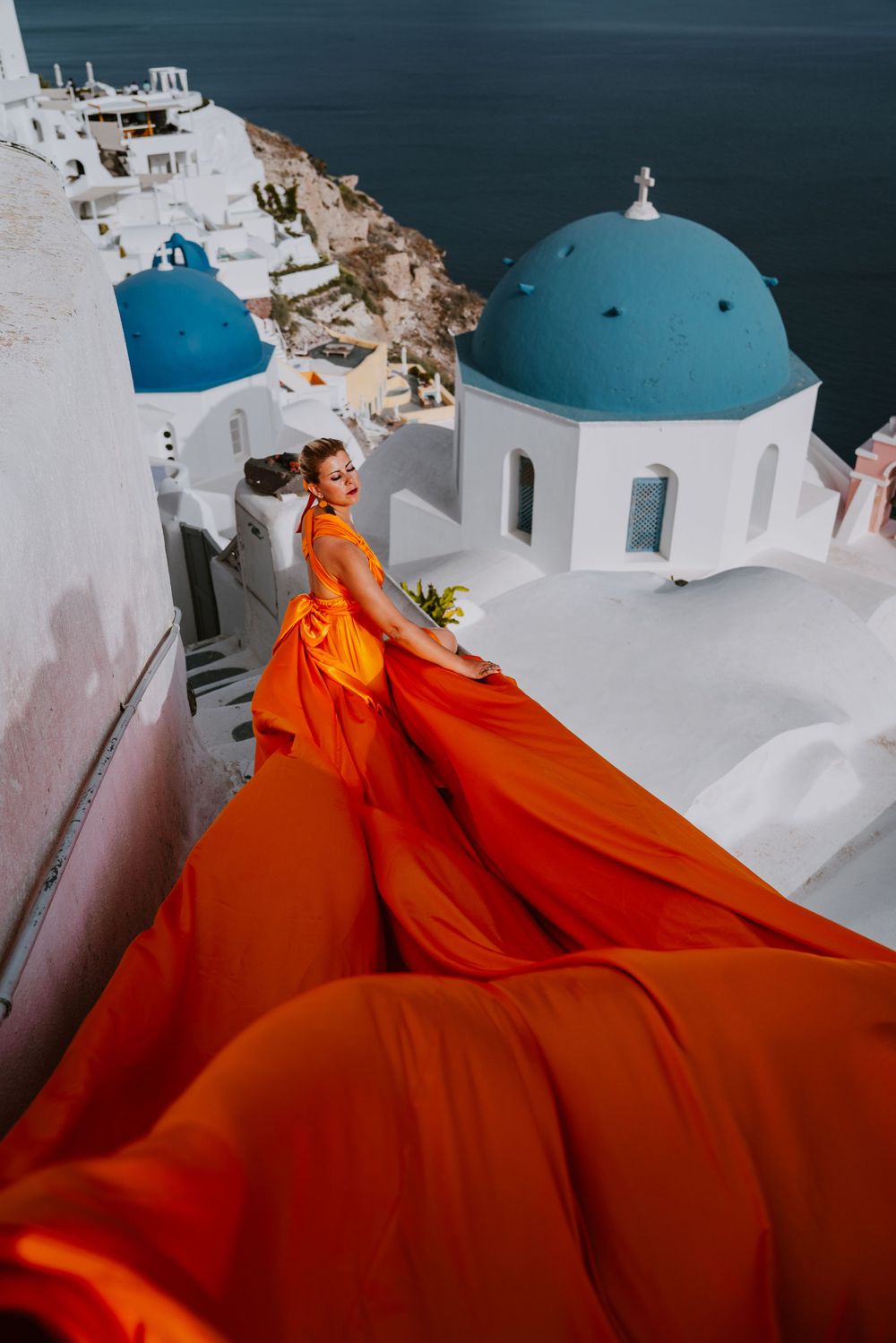 Orange flowing dress fabric creates dramatic contrast against iconic blue domed church in Santorini.