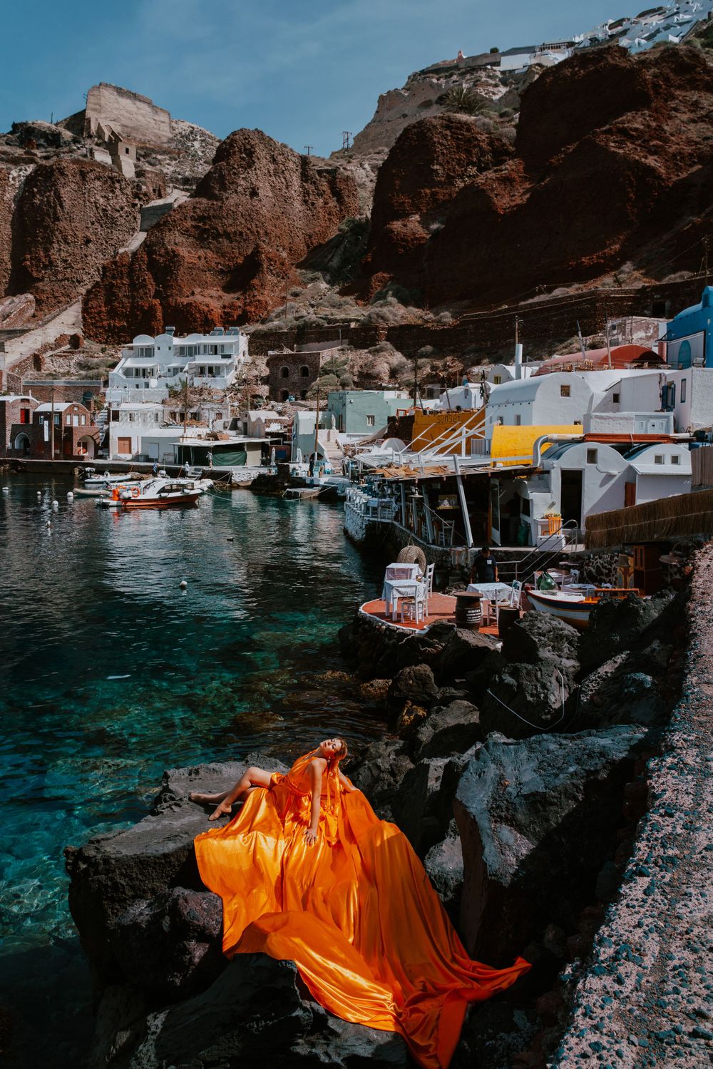 Boats dock in a Mediterranean harbor with orange fabric draped over coastal rocks.