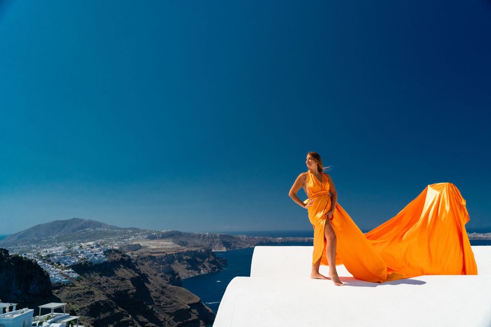 Orange fabric billows dramatically on white rooftop overlooking Mediterranean landscape.
