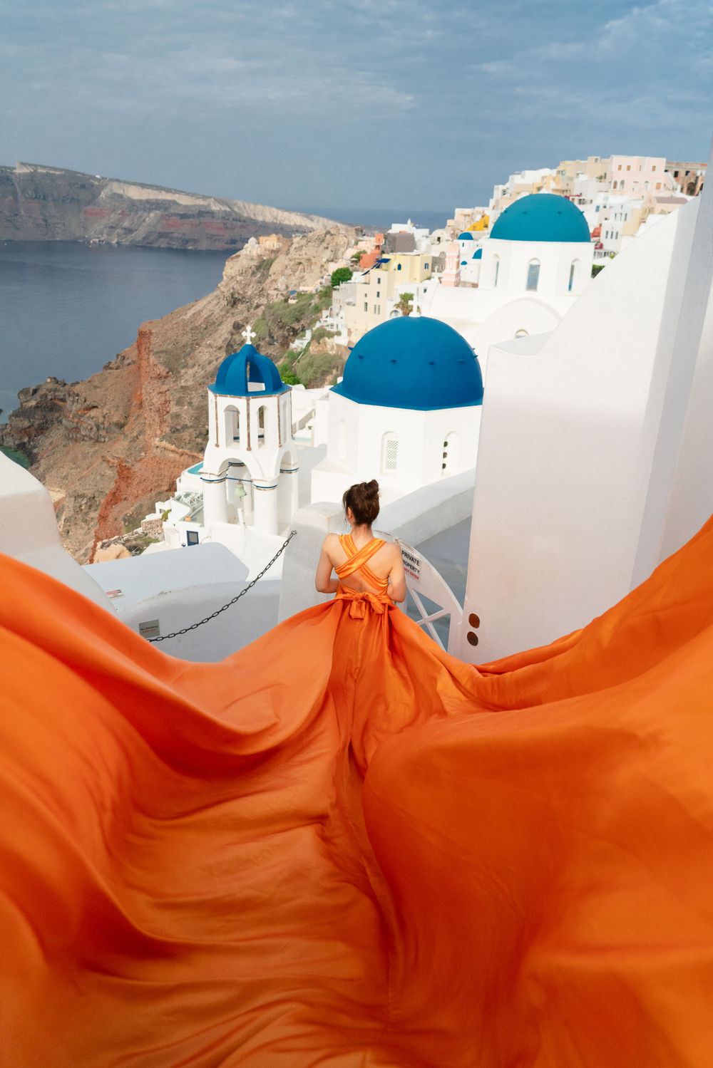 A flowing orange dress billows dramatically against iconic white buildings and blue domes of Santorini, Greece.
