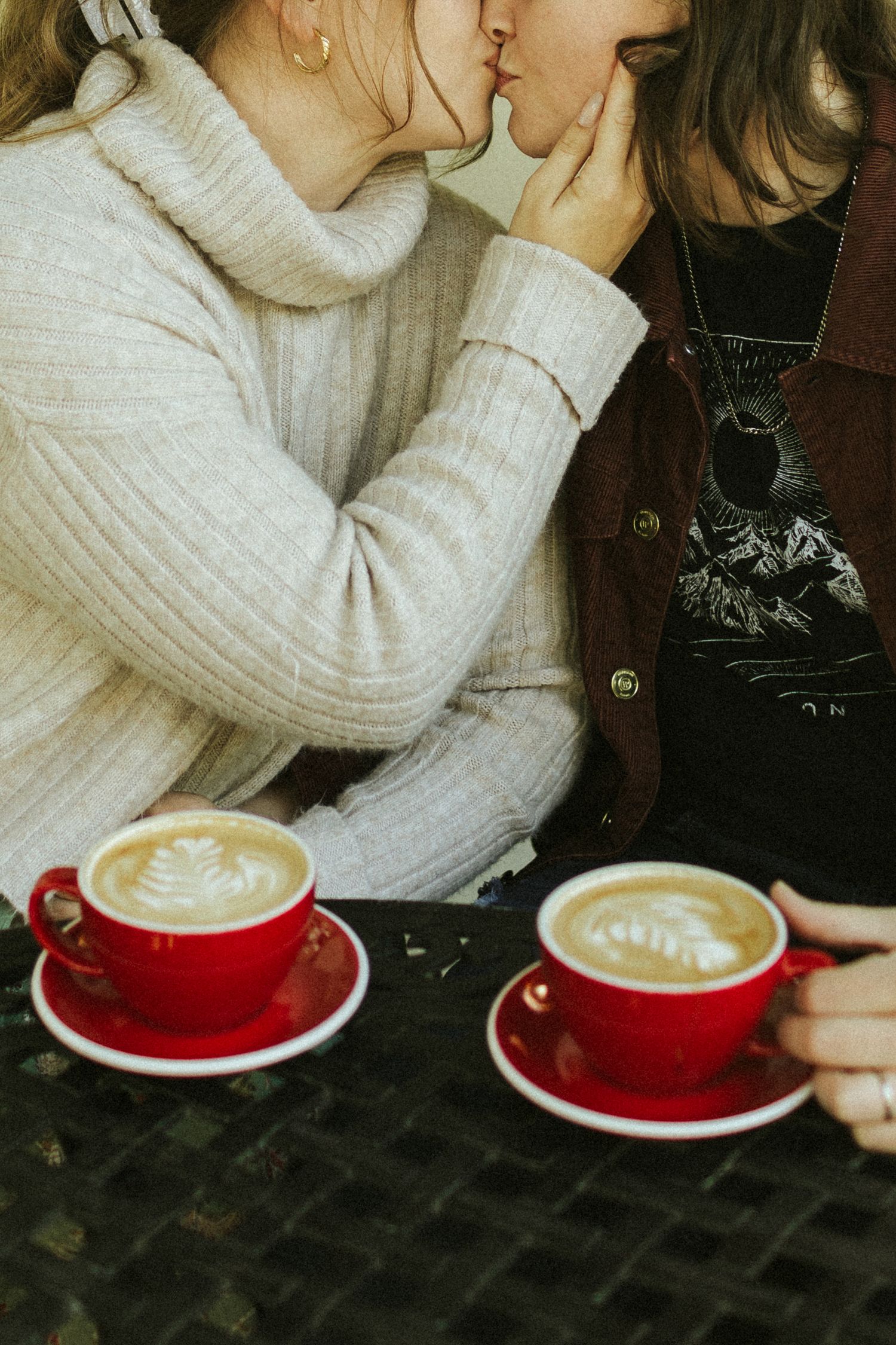 A close-up image of two women kissing behind red coffee mugs with lattes