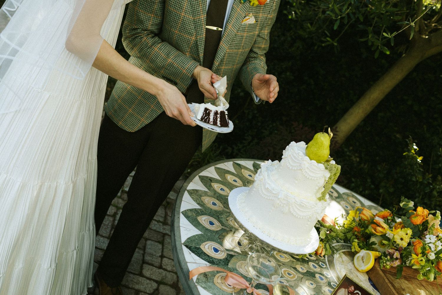 A bride and groom standing side by side across from their wedding cake, sliding a piece of it onto their plate