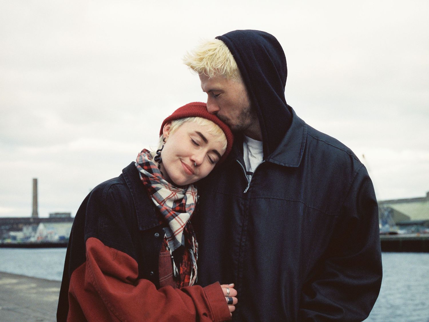 A couple in winter coats share an intimate moment near a waterfront on an overcast day.