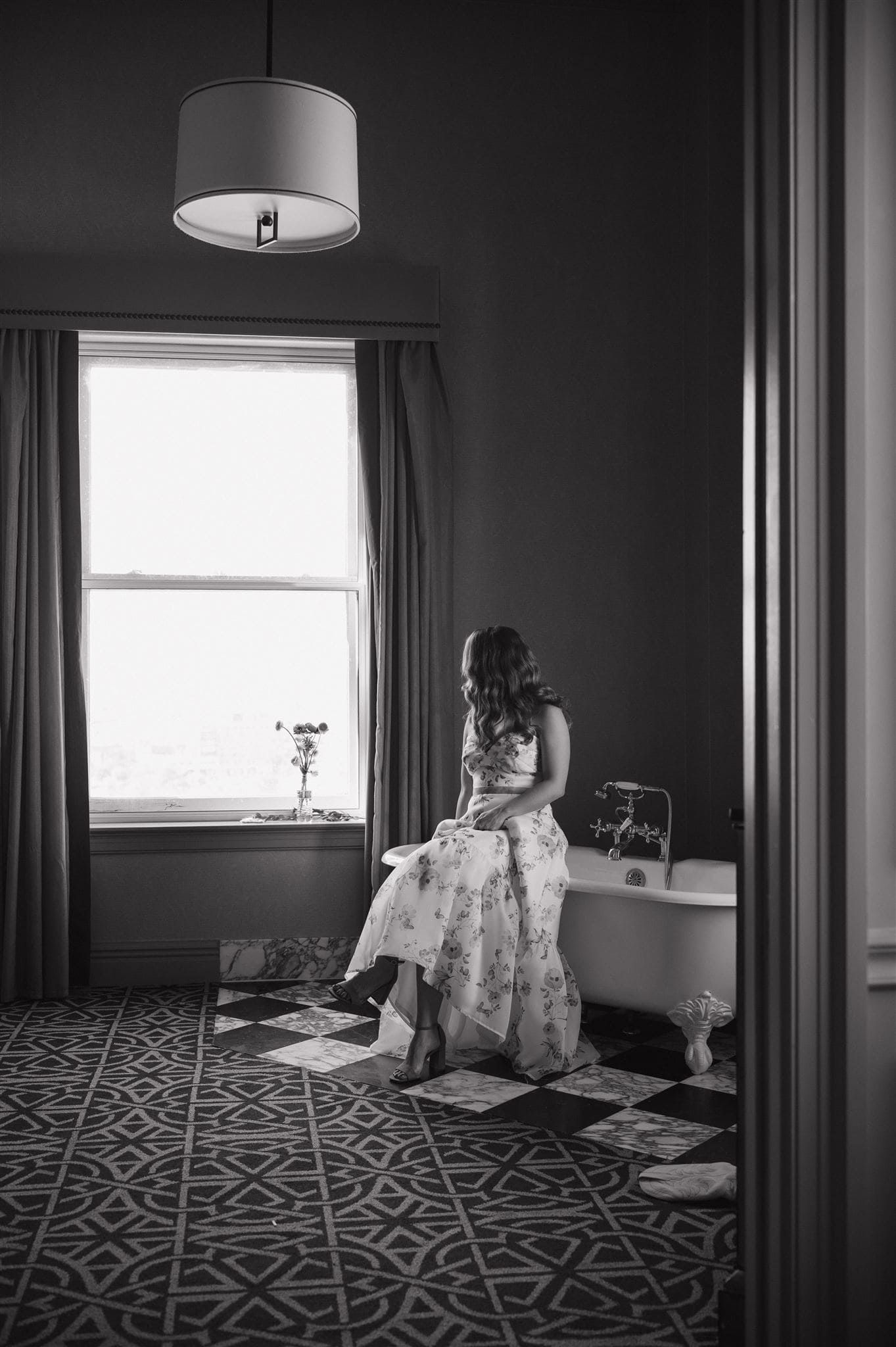 Black and white photo of a figure sitting by a window in an elegant room with patterned floors.