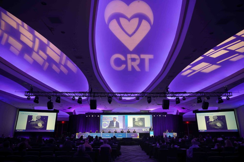 Purple-lit conference room with multiple screens showing video presentations and a large audience in attendance.