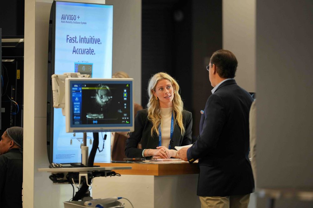 A professional conversation takes place at a display booth with a medical ultrasound monitor visible in the background.