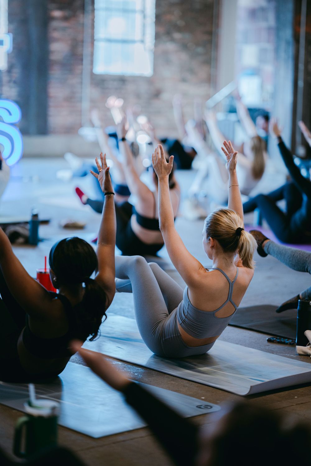 Yoga class participants lie on mats with raised arms during a relaxing stretching exercise in a sunlit studio.