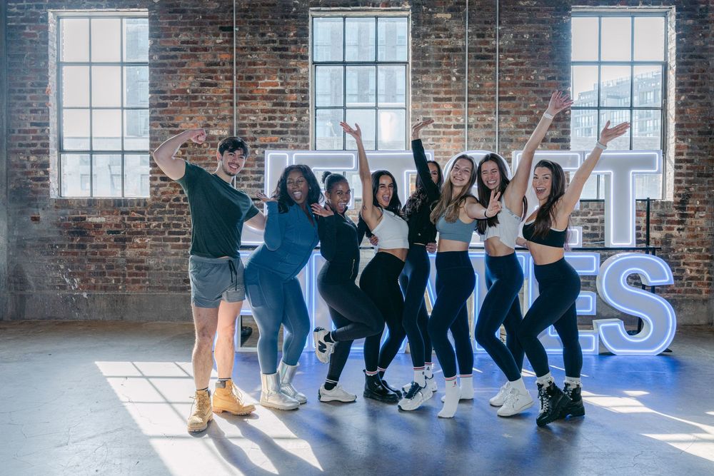 A group of dancers in athletic wear strike energetic poses together in a sunlit industrial studio space with brick walls.