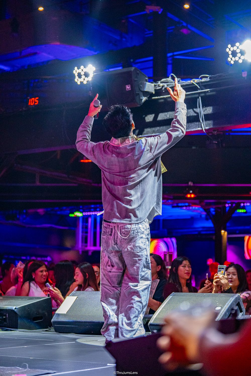 Performer in silver jumpsuit energetically entertains crowd at nightclub with dramatic gestures under blue lighting.