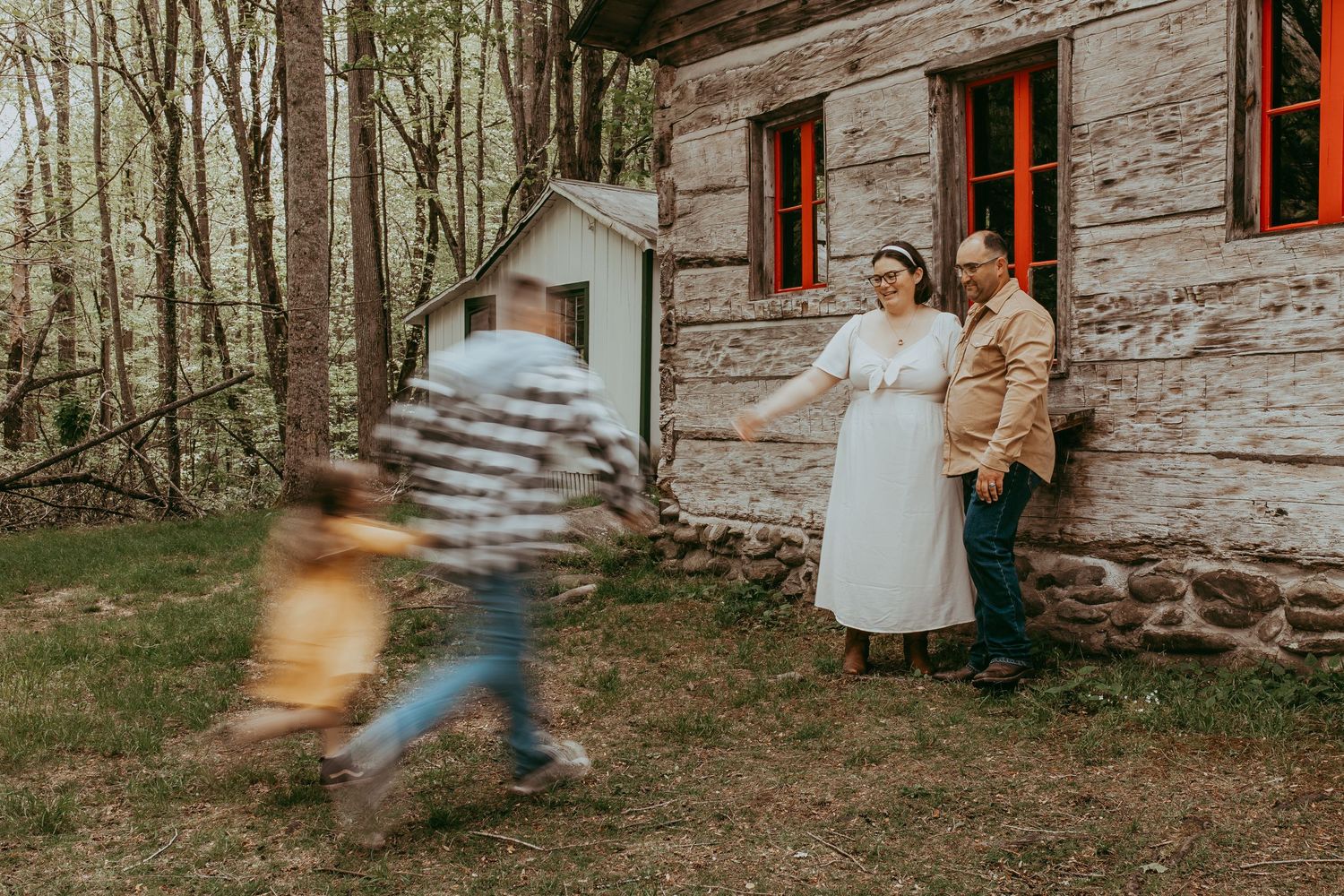 Family photo with couple leaning against rustic log cabin while two children run toward them outdoors. Smokey Mountains, Tennessee