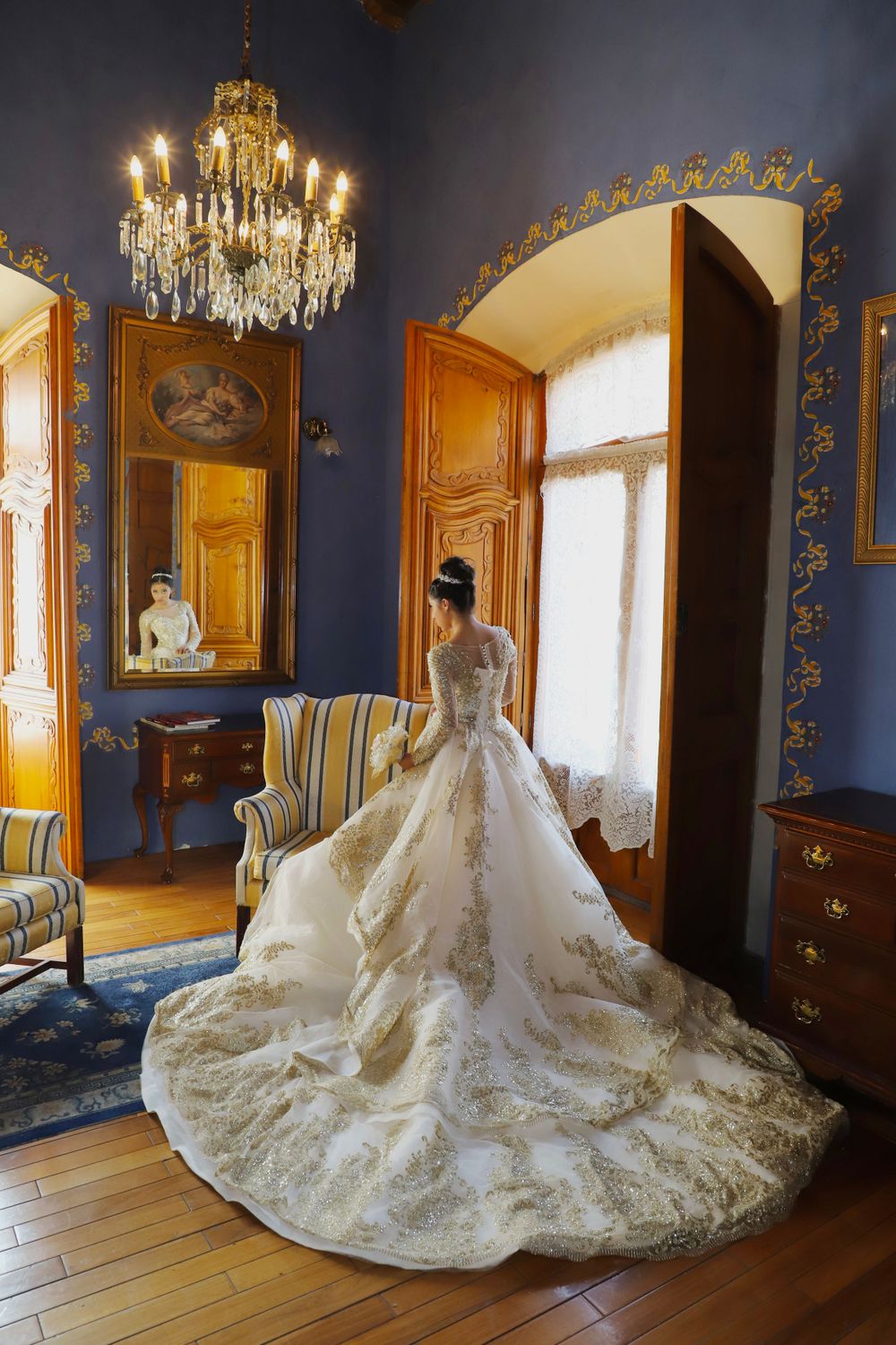 Elegant white wedding dress with long train displayed in blue room with chandeliers.