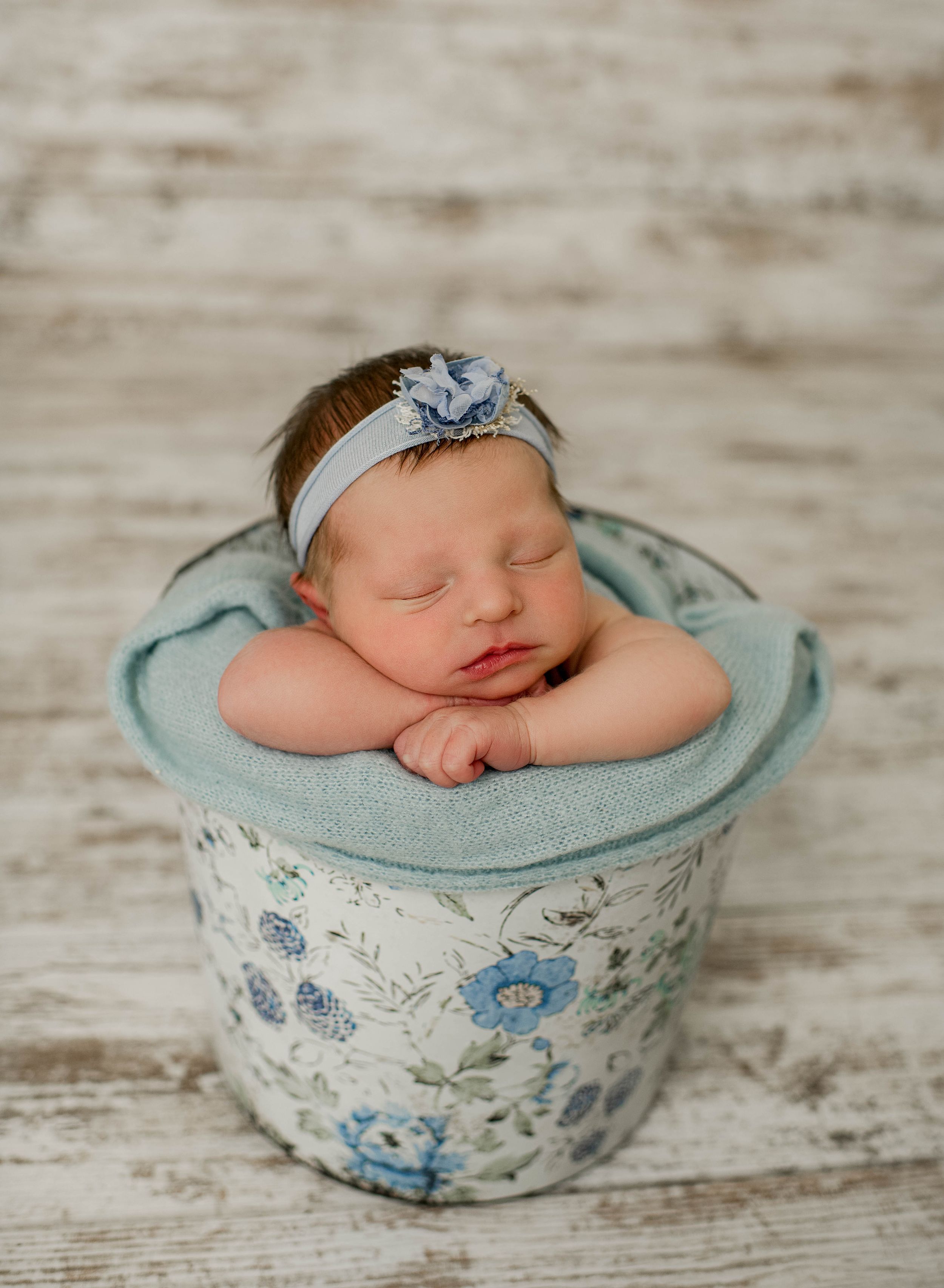 Sleeping newborn nestled in a vintage blue and white floral bucket on rustic wooden background.