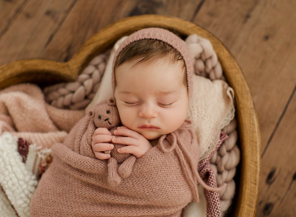 Newborn baby sleeping peacefully wrapped in soft brown swaddle blanket in wooden basket.