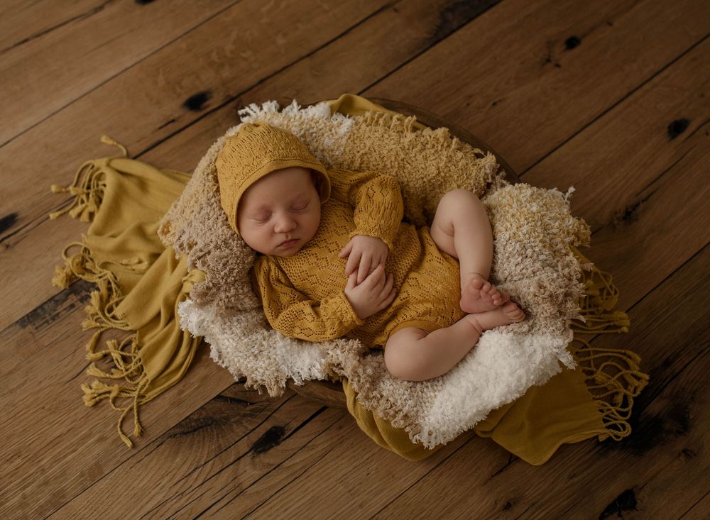 Sleeping baby in yellow knit outfit curled up on cream blanket against wooden floor background.