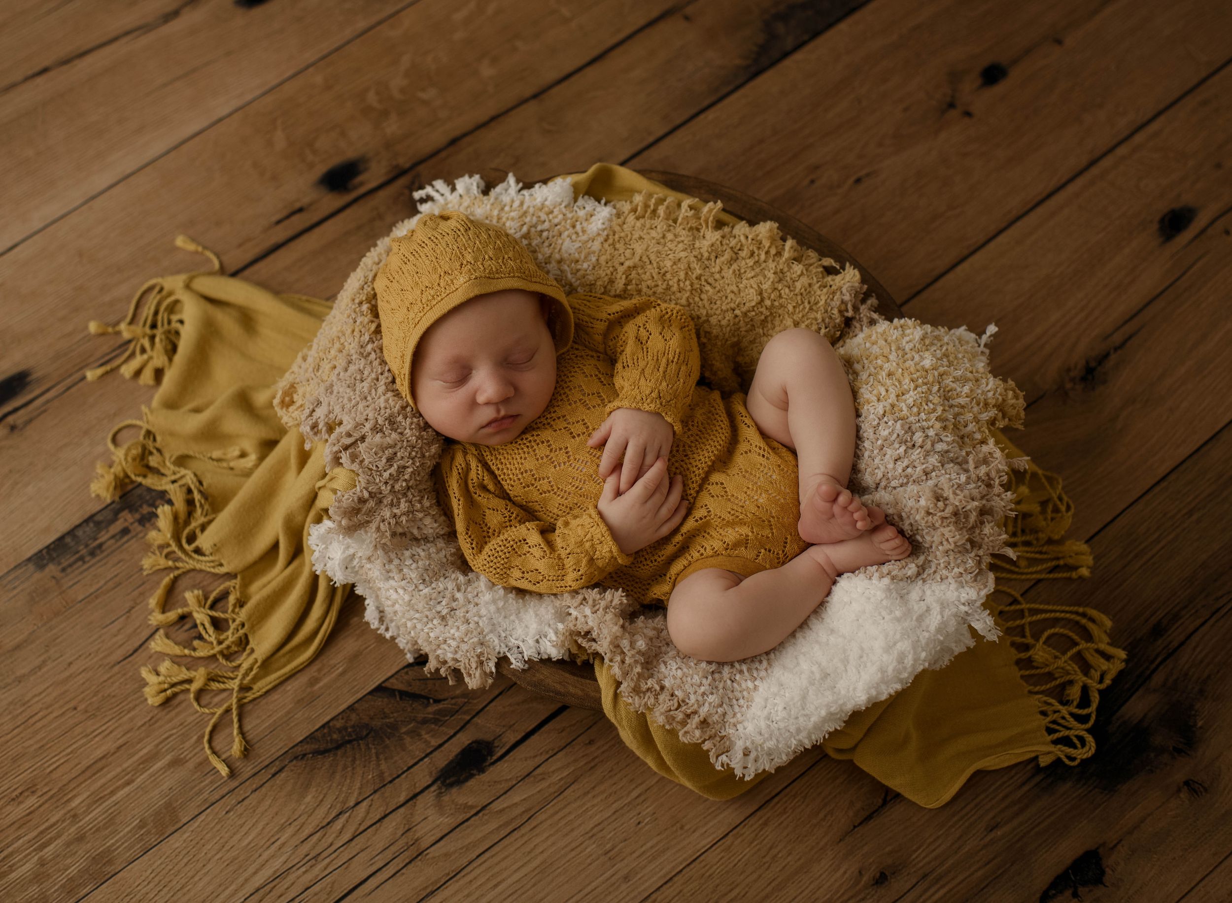 Sleeping baby in yellow knit outfit curled up on cream blanket against wooden floor background.