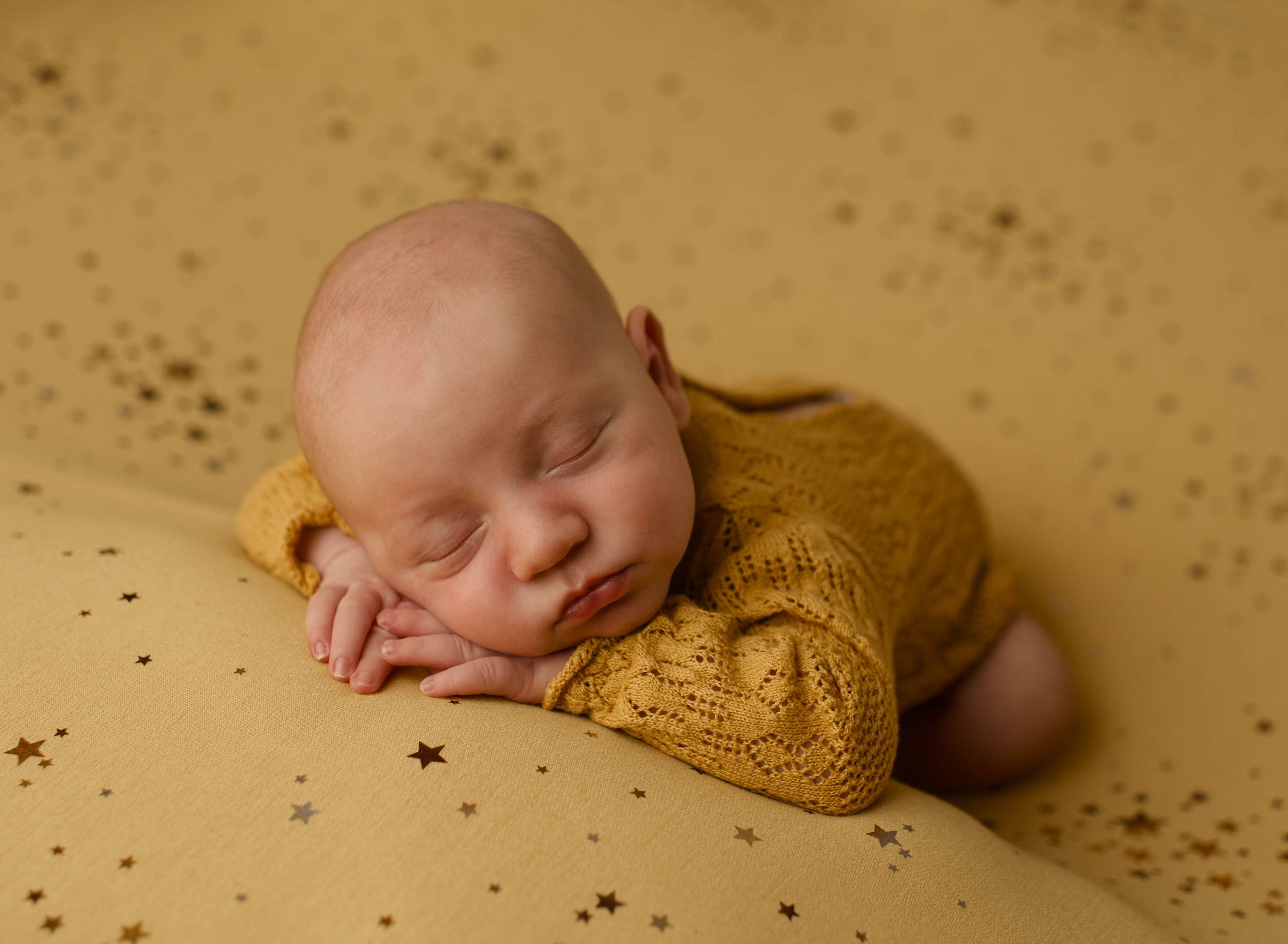 A sleeping newborn wearing a mustard yellow knit wrap lies on a textured beige backdrop.