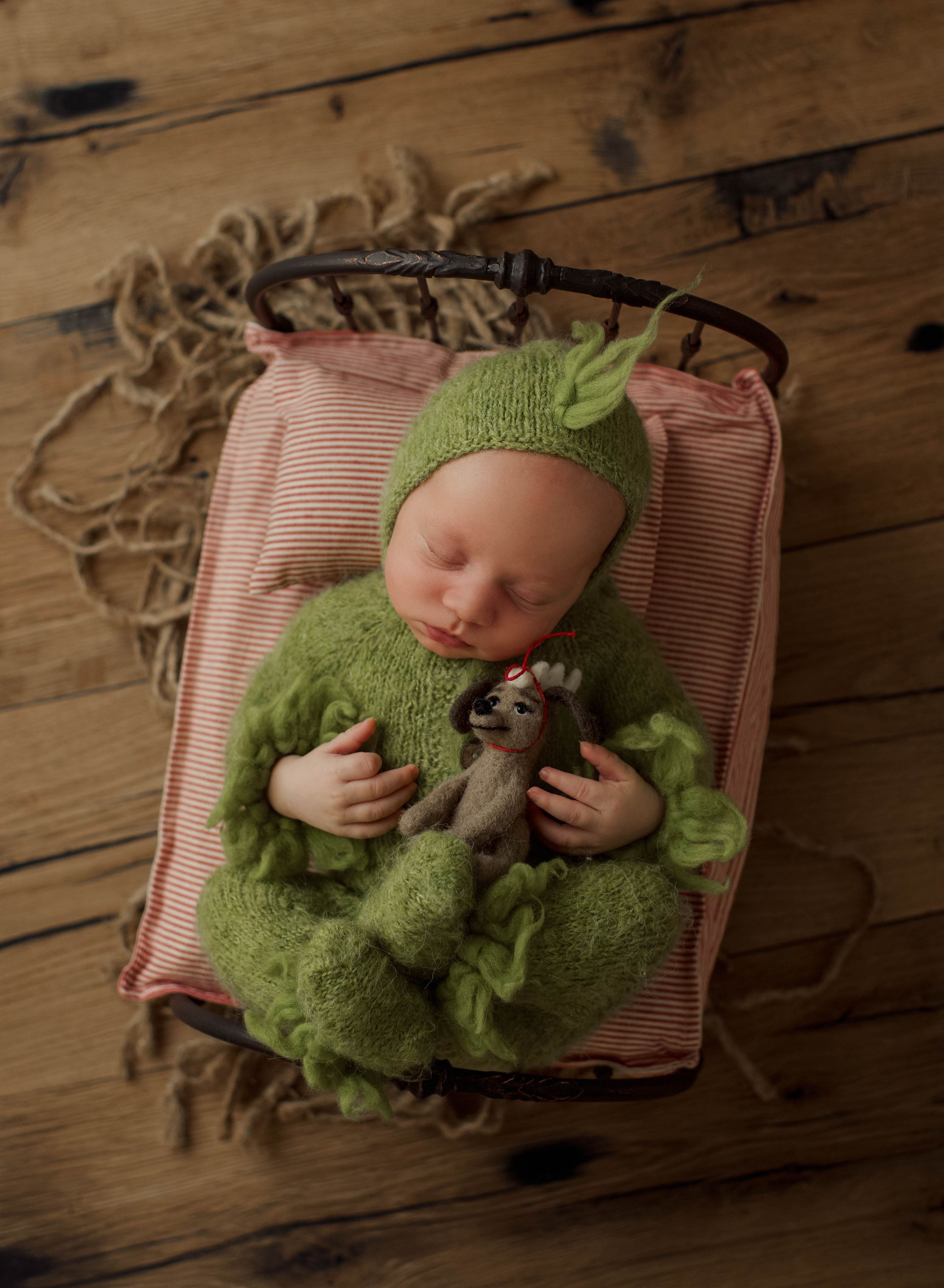 A small baby wearing a green knit outfit lies sleeping on a pink and white striped cushion on wooden flooring.