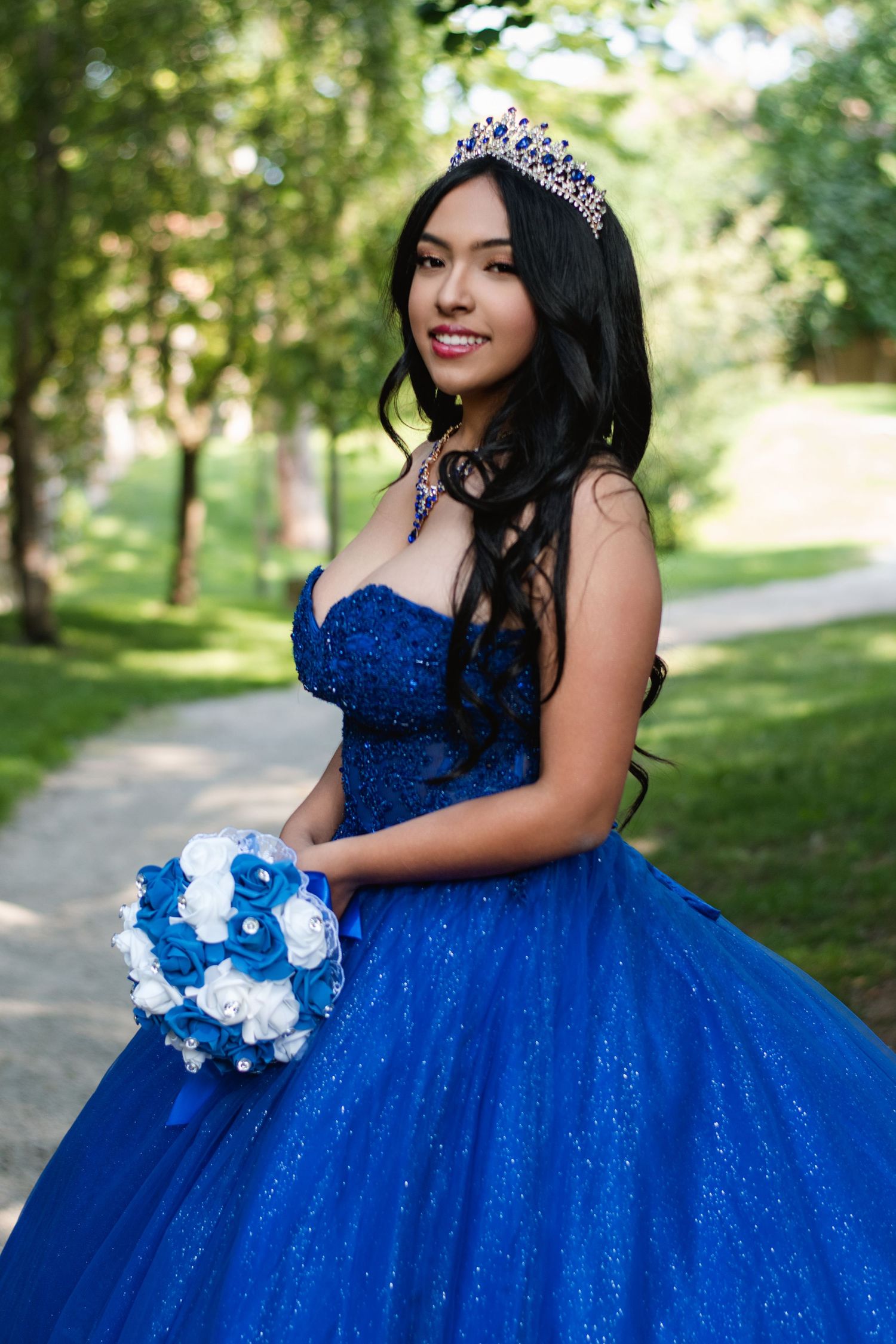 Young woman in stunning blue quinceañera ball gown with crown holds bouquet in park. Toronto GTA