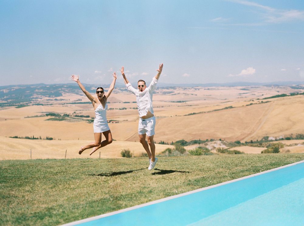 Newlyweds happily leaping by a pool at a white wedding pool party in Tuscany, Italy.