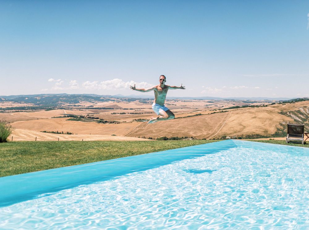 A groom joyfully jumping into the pool at a white wedding pool party in Tuscany, Italy.