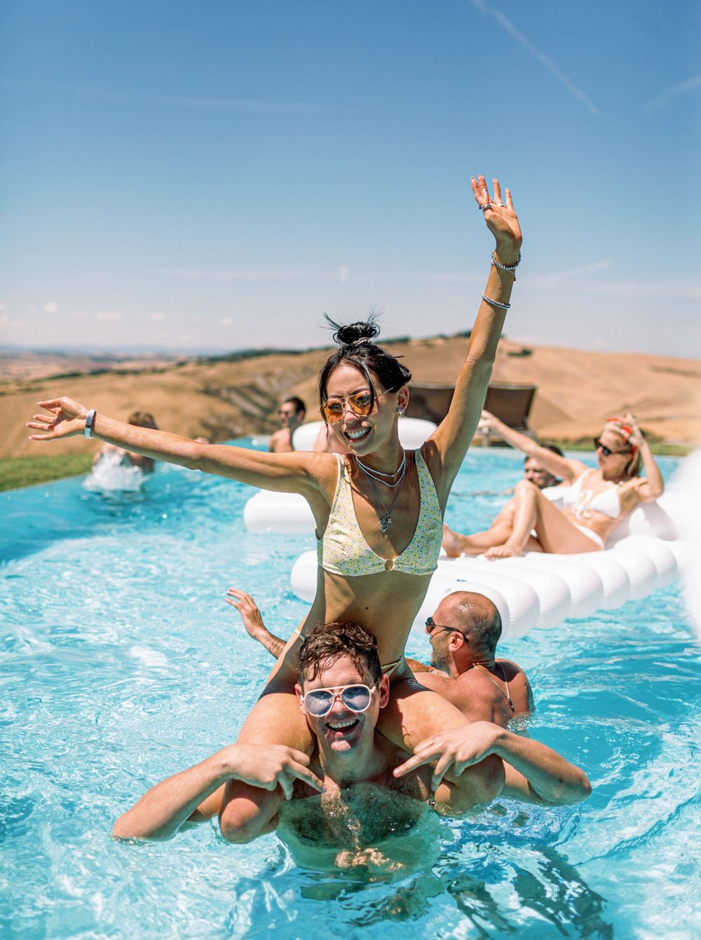 A woman in a bikini joyfully rides on a man in a pool at a white wedding pool party in Tuscany, Italy.