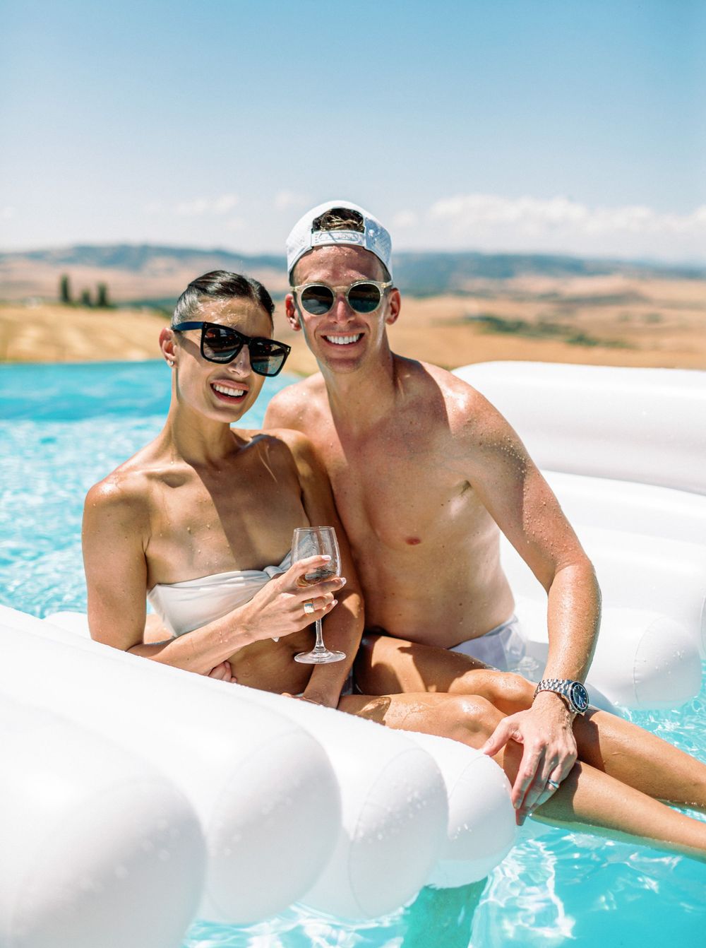 A couple in a pool, holding wine glasses, at a white wedding pool party in La Bandita Countryhouse, Tuscany, Italy.