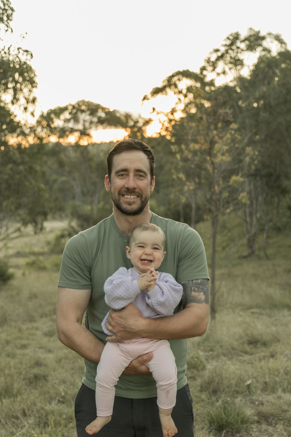 A tender moment at sunset shows a parent holding an infant in a green outdoor setting.