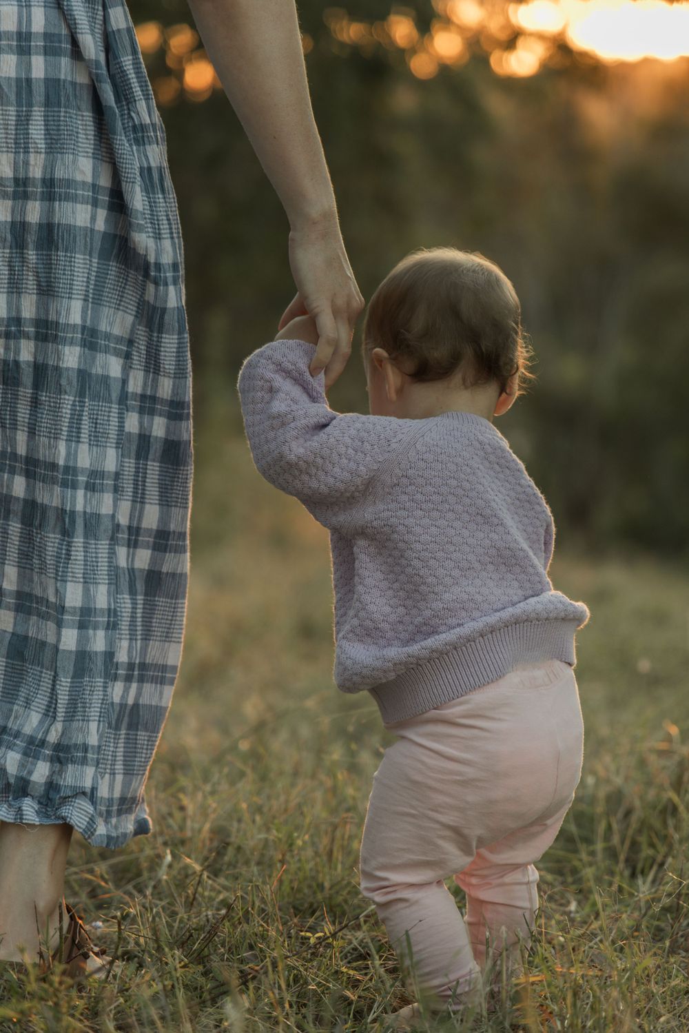 A small child in pink pants holds onto a parent's hand while learning to walk in a grassy field at sunset.