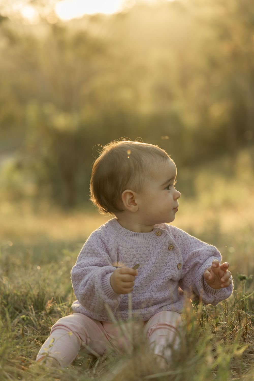 Baby in sweater sitting in sunlit grass during golden hour.