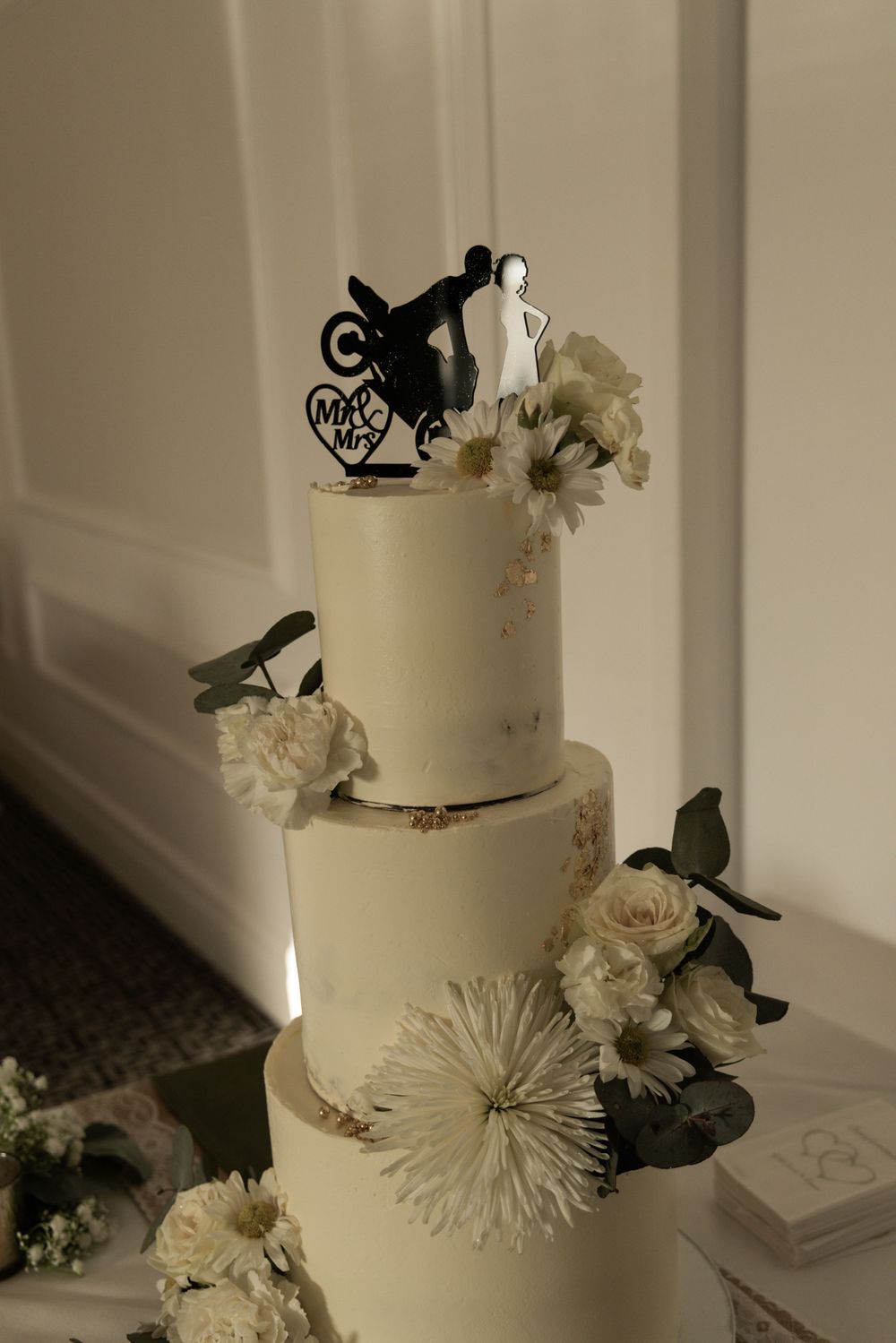 Elegant white tiered wedding cake decorated with flowers and motorcycle couple topper.