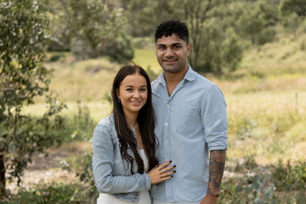 A couple poses together outdoors in casual attire against a natural landscape backdrop.