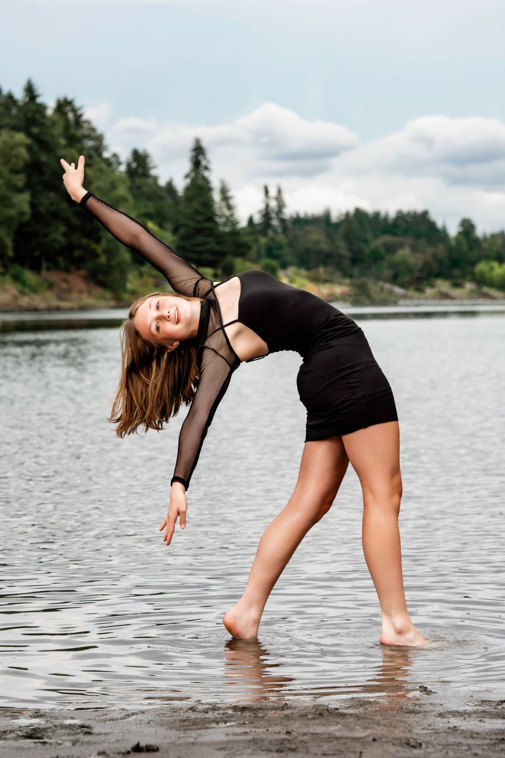 Dancer in black dress performs graceful pose while standing in shallow lake water with evergreen forest backdrop.