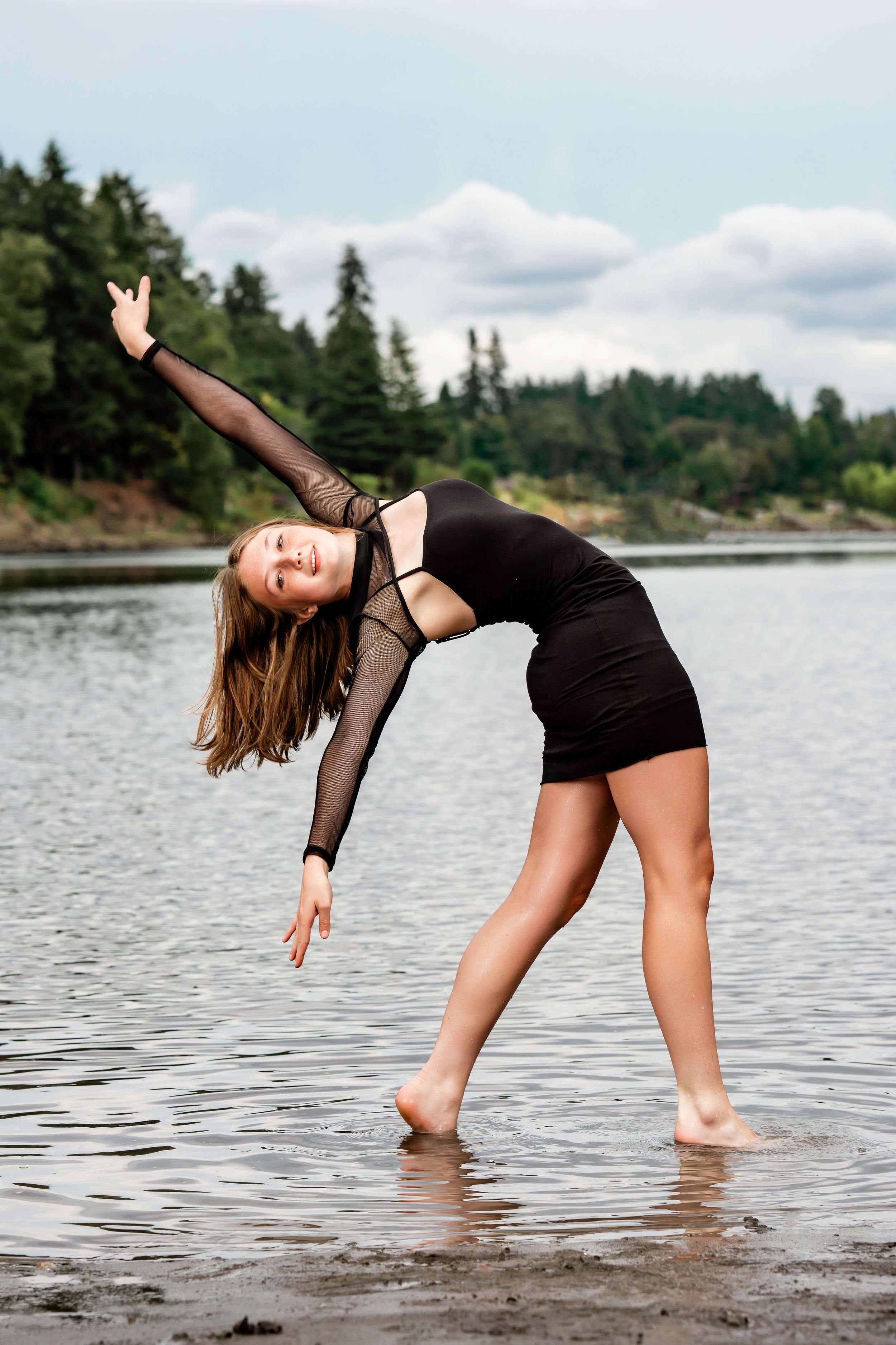 Dancer in black dress performs graceful pose while standing in shallow lake water with evergreen forest backdrop.
