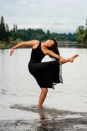 Dancer in black dress performs graceful pose while standing in shallow lake water with evergreen forest backdrop.