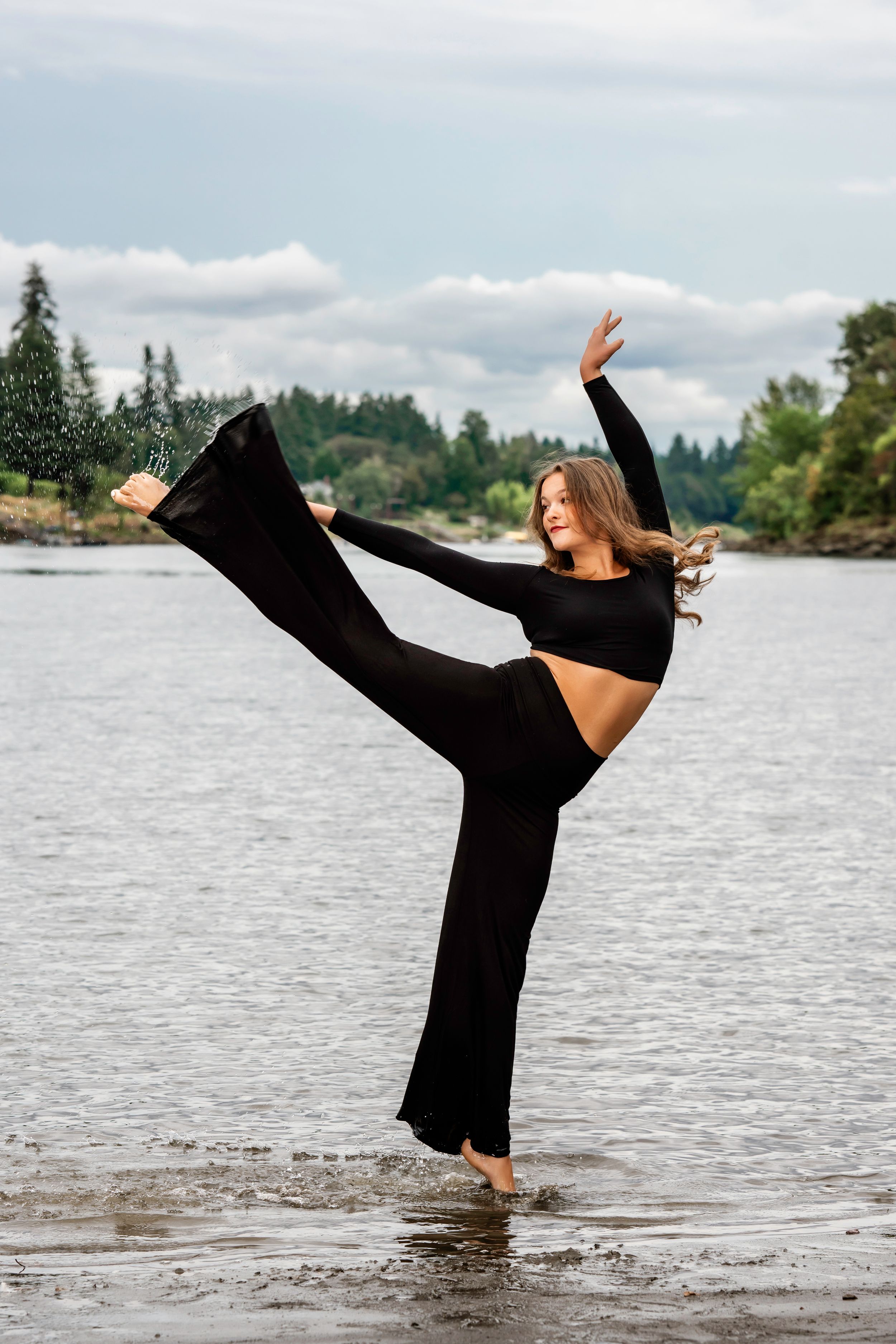 Dancer in black outfit performs high leg kick pose while standing in shallow lake water with forest backdrop. High School Dance Team photos.