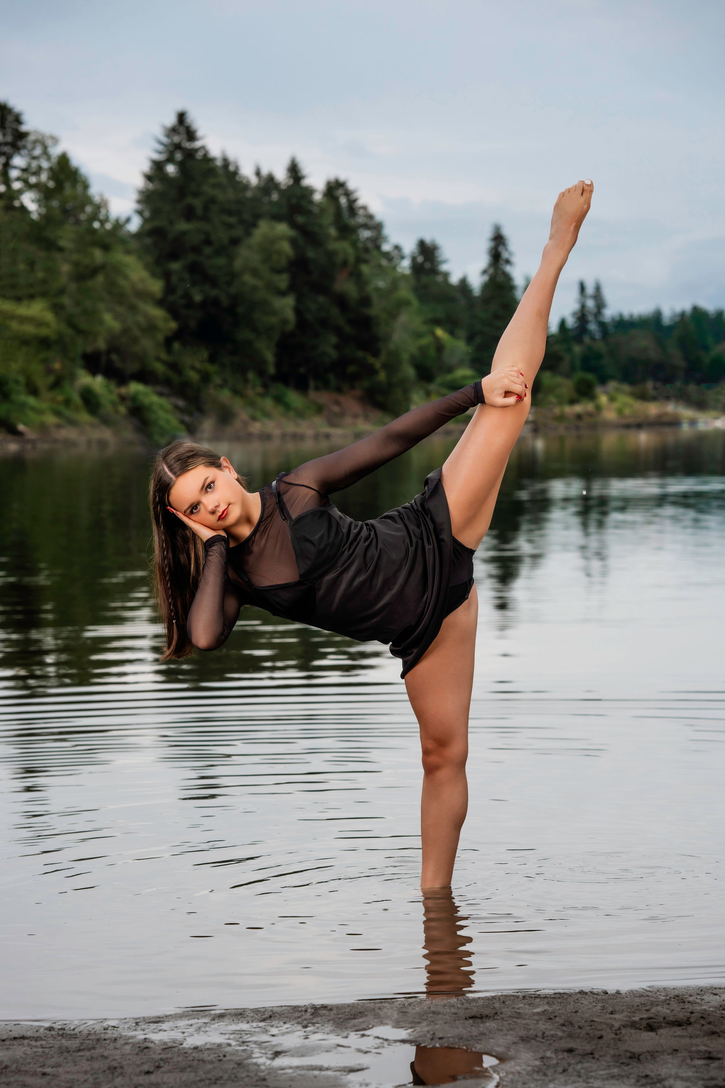 A Lakeridge High School Dancer in a black leotard performs a high kick pose while standing in shallow water near a forested riverbank.