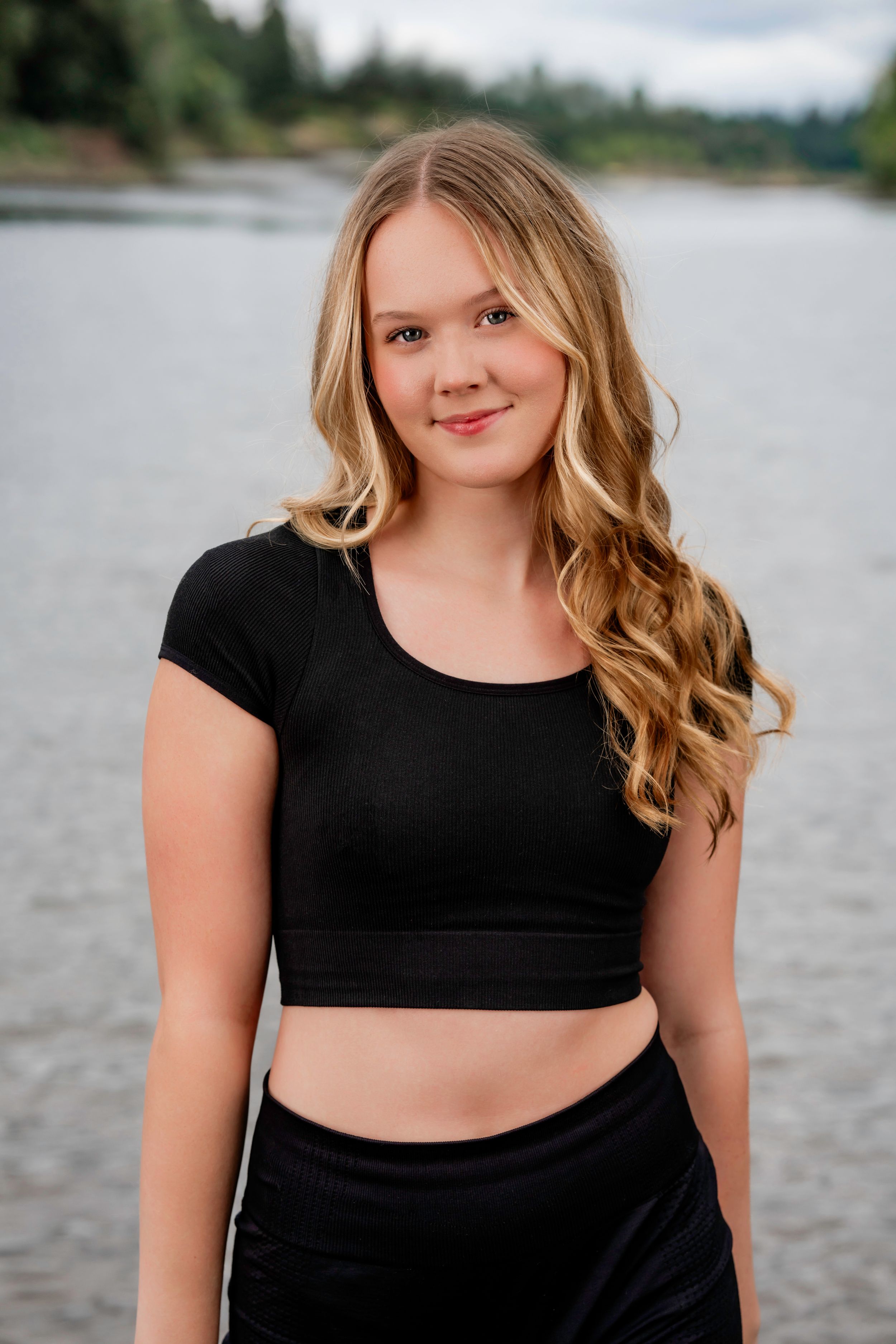 Lakeridge High School Dance Team dancer wearing a black crop top and dark pants poses outdoors near a lake with a forest backdrop.