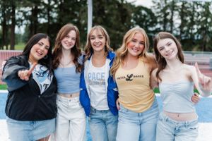Five dance team dancers in casual clothing including denim jeans and t-shirts pose together outdoors on a sunny day.