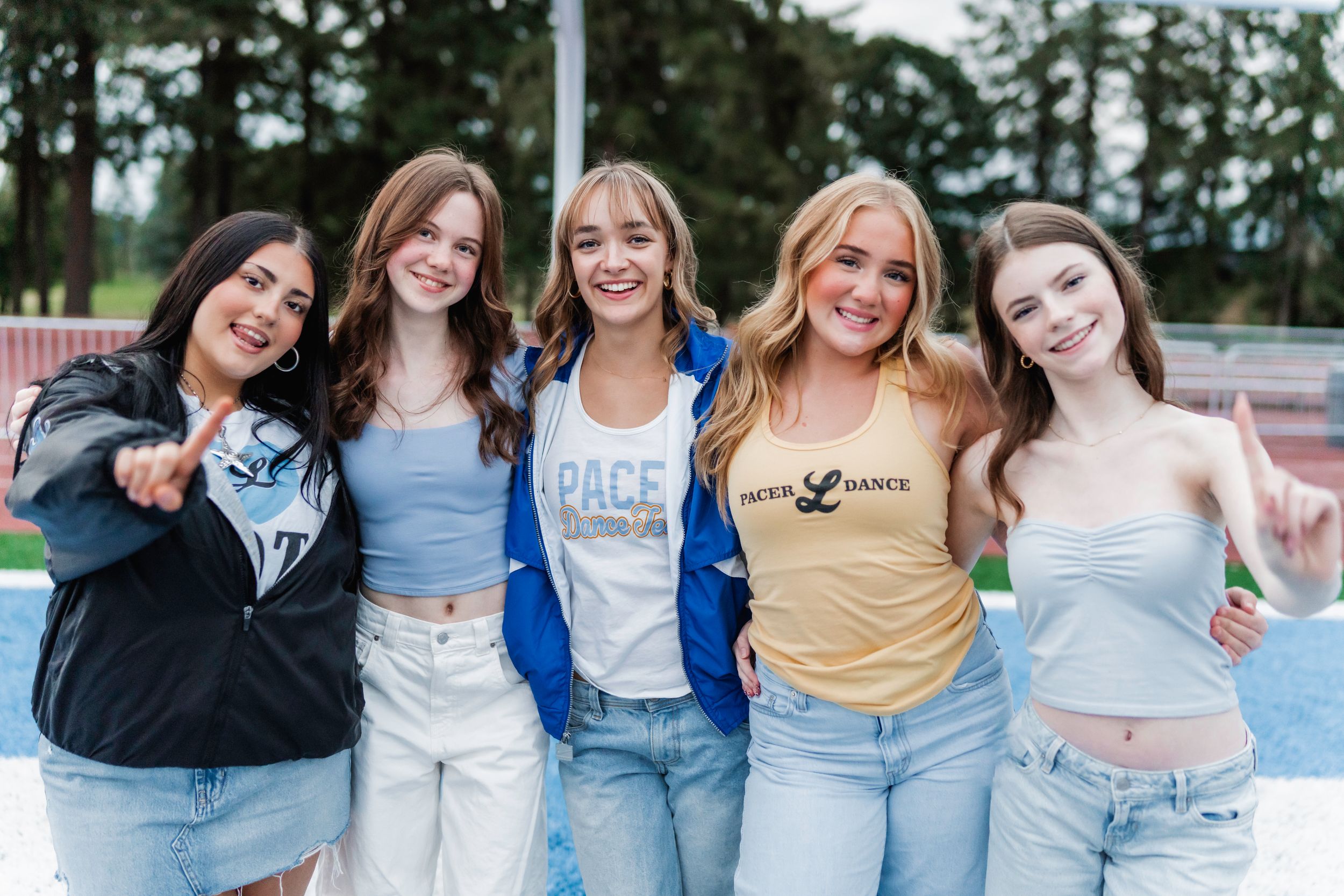 Five dance team dancers in casual clothing including denim jeans and t-shirts pose together outdoors on a sunny day.