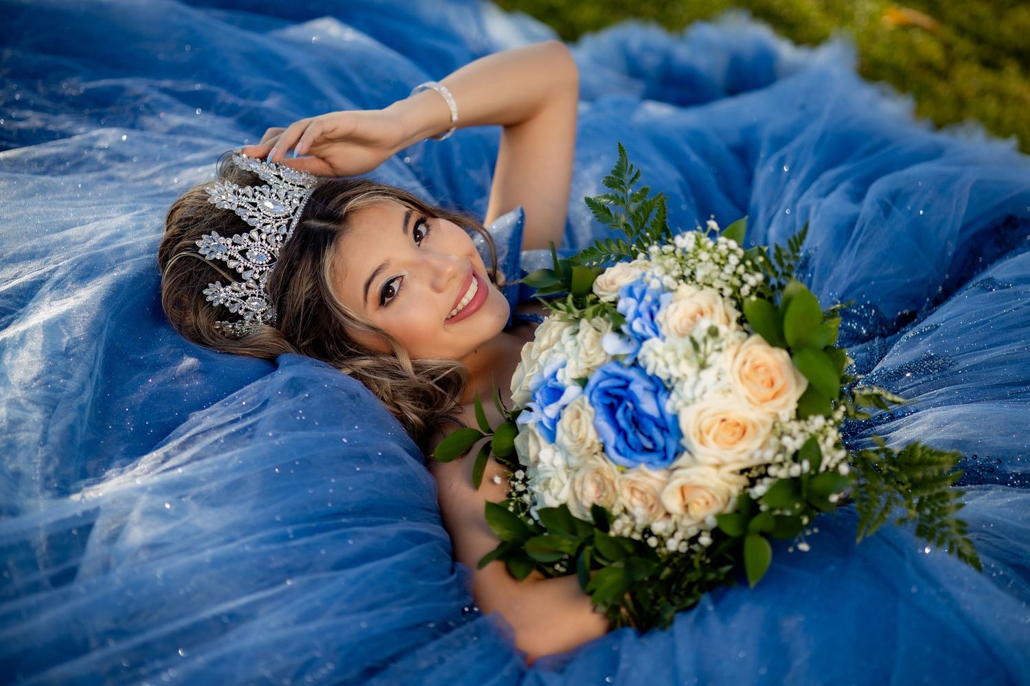 A smiling quinceañera in a blue ball gown and tiara poses lying down holding a bouquet of roses.