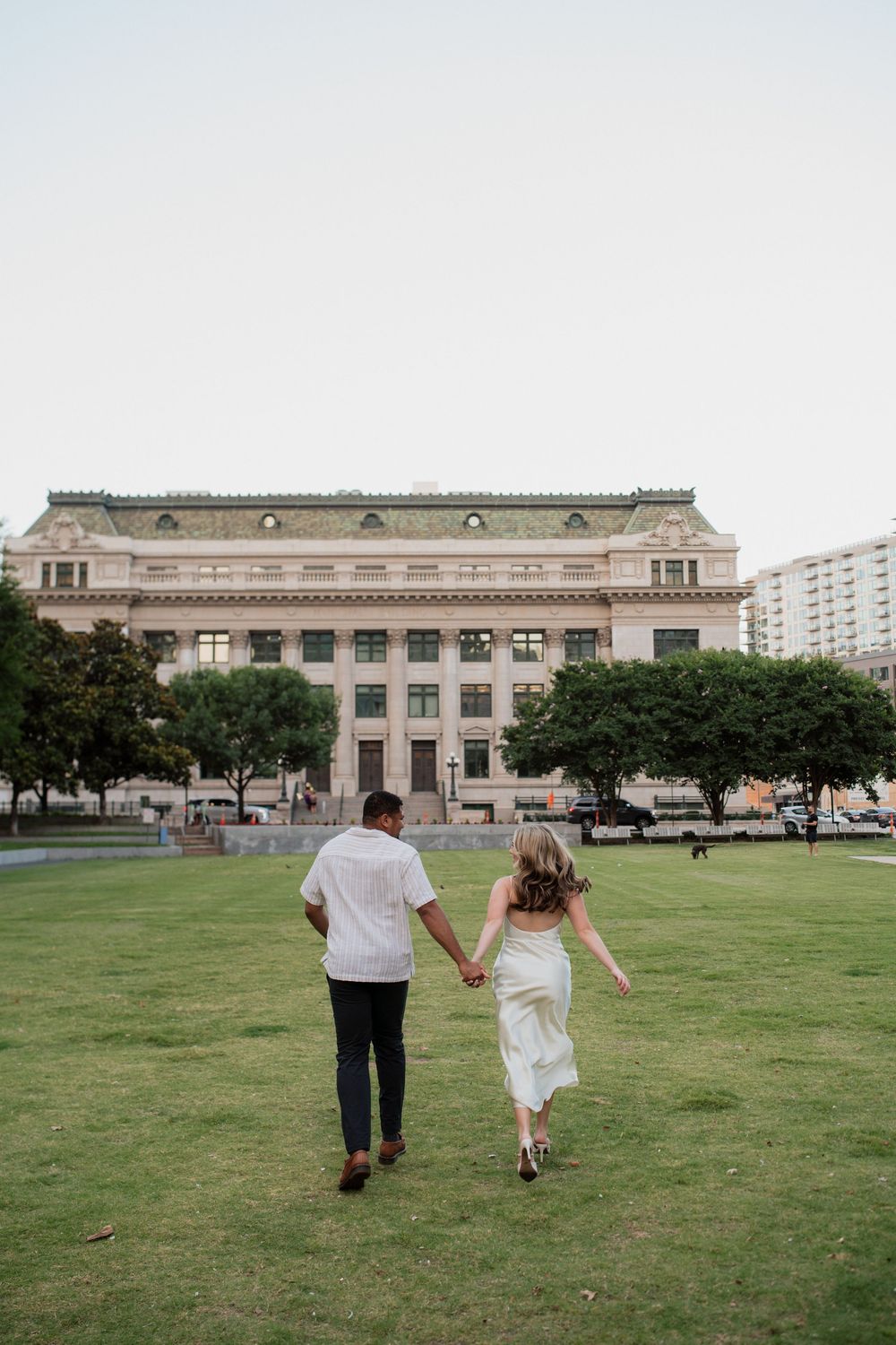 A couple walks away holding hands on a green lawn towards a historic stone building on a cloudy day.