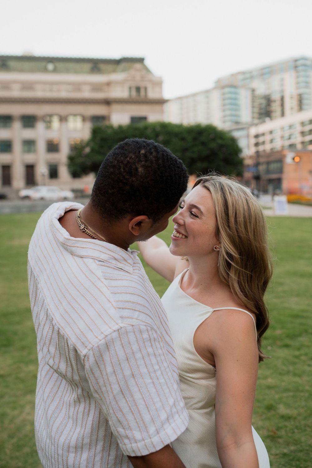 Happy couple in white embracing on a lawn with urban buildings in the background.