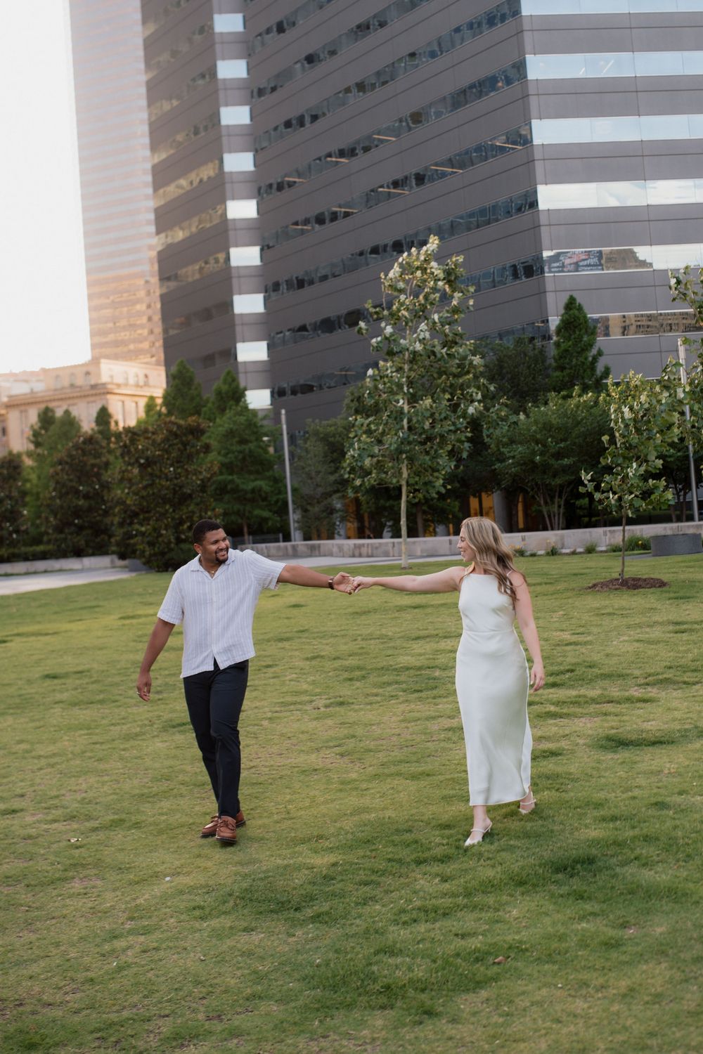 A couple in white and navy outfits playfully runs across a grassy lawn with tall buildings in the background.