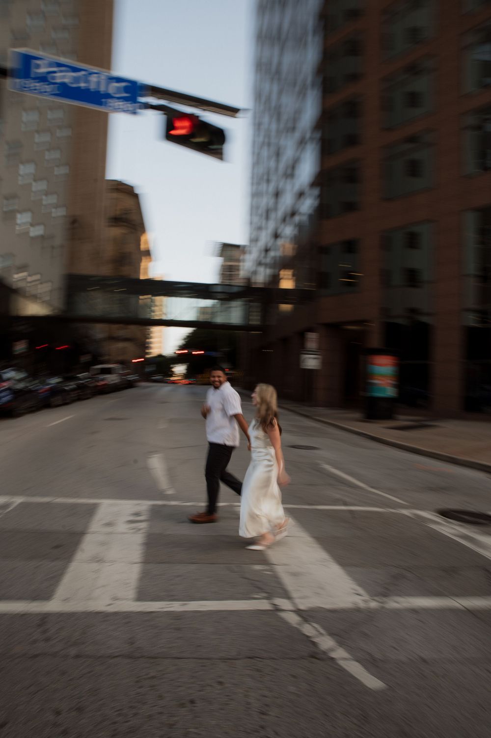 A couple walks across an urban street intersection with tall buildings and a street sign in motion blur.